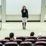 Female speaker presenting to an audience in a modern auditorium setting.